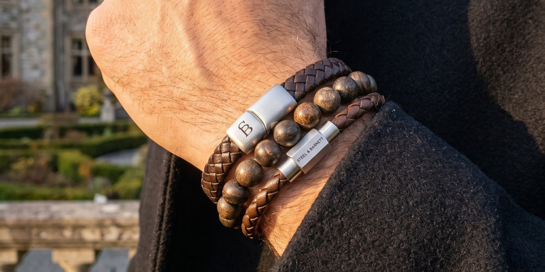 A close-up of a person's wrist wearing multiple leather and wood bracelets, including braided brown leather, round wooden beads, and a silver-toned magnetic band with a white logo plaque, set against a blurred outdoor background.