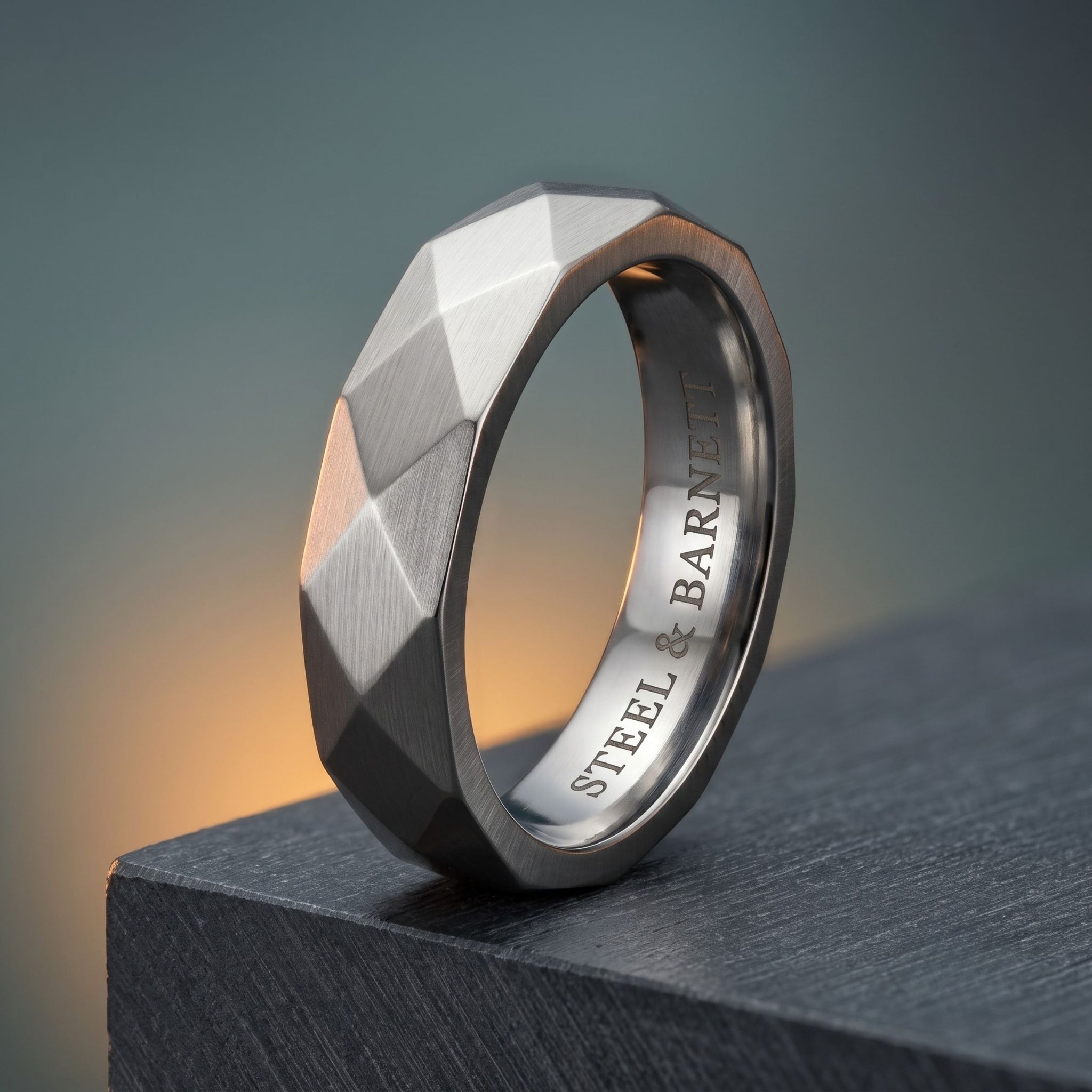 A faceted geometric steel ring with a brushed silver exterior and a polished inner band engraved with 'STEEL & BARNETT', resting on a dark textured block against a soft gray background.