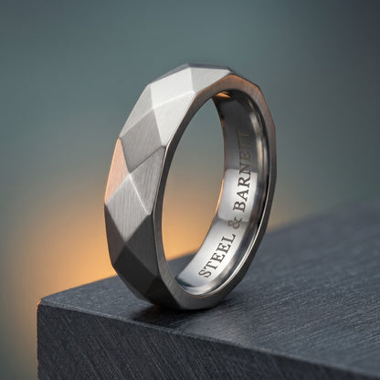 A faceted geometric steel ring with a brushed silver exterior and a polished inner band engraved with 'STEEL & BARNETT', resting on a dark textured block against a soft gray background.