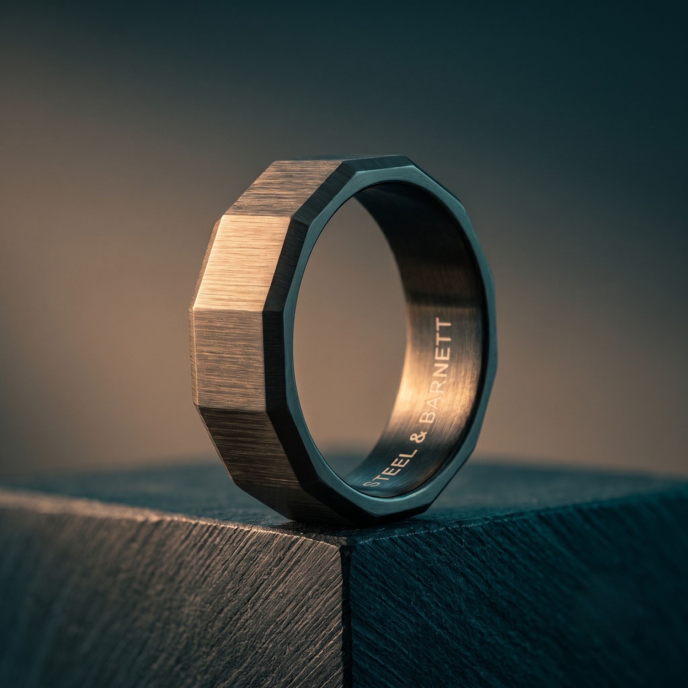 A geometric octagonal ring with a brushed bronze-toned outer surface and a dark, polished inner band engraved with 'STEEL & BARNETT', resting on a dark textured surface.