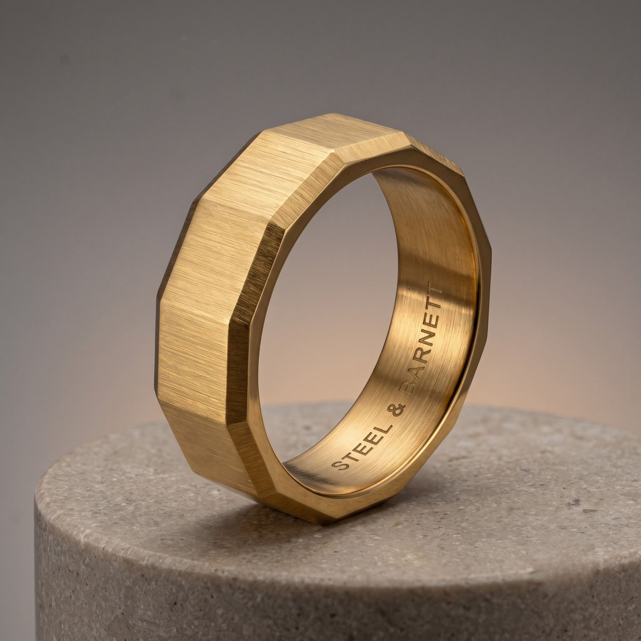 Gold stainless steel men's ring with a brushed geometric design displayed upright on a light stone pedestal