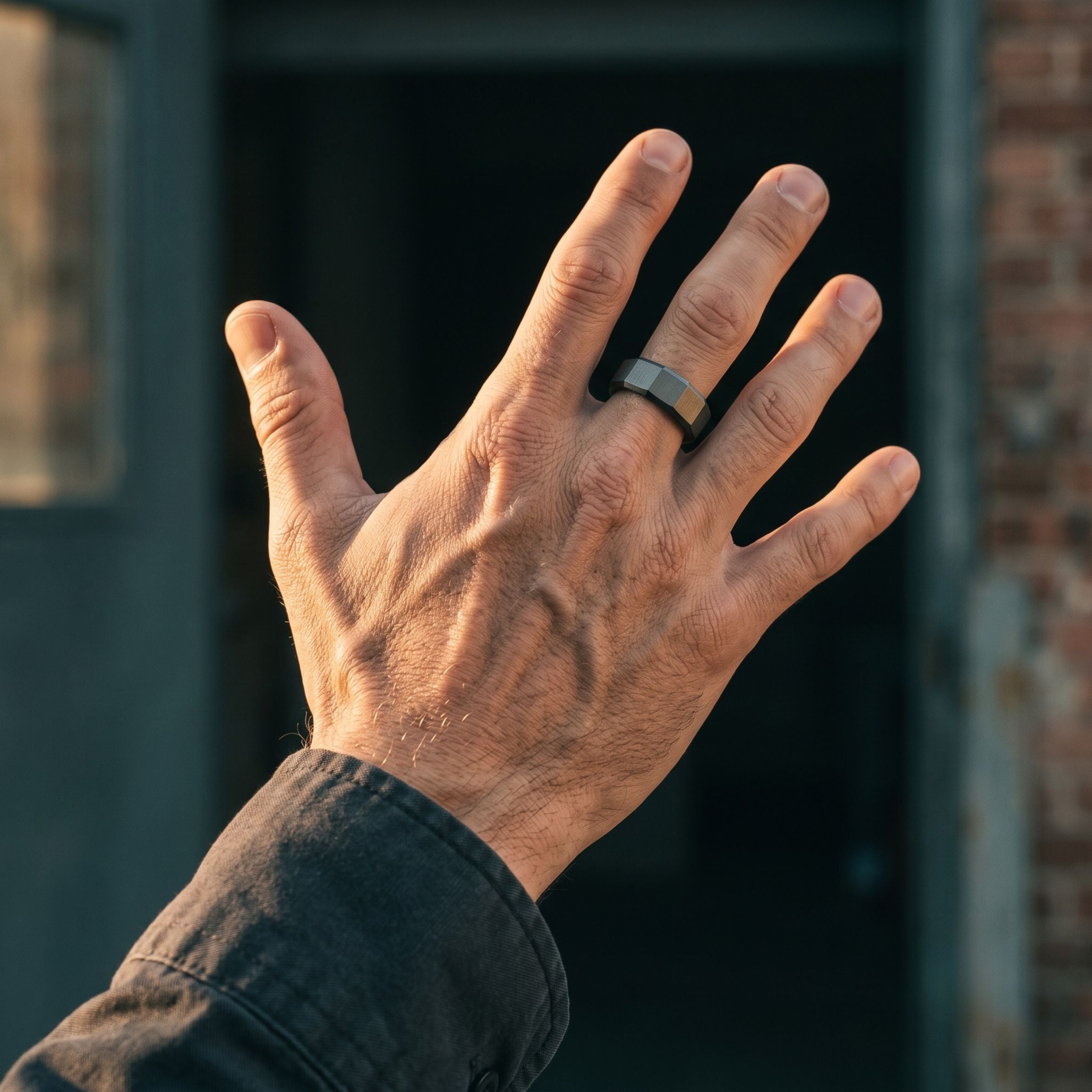 A close-up of a person's hand wearing a dark metallic band ring, set against a blurred background of a dark doorway and brick wall.