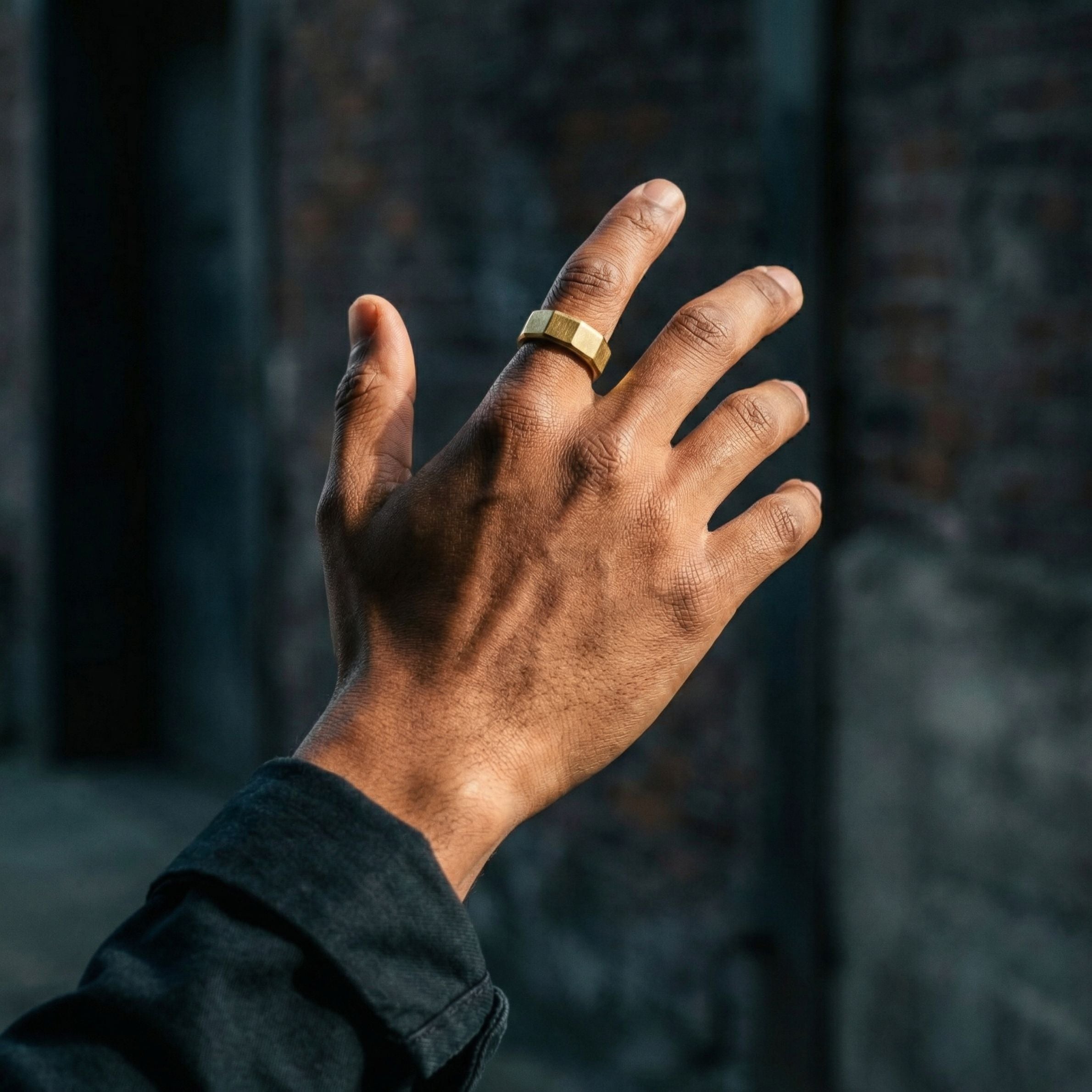 Gold stainless steel men's ring with a brushed geometric design worn on a raised hand in an outdoor industrial setting
