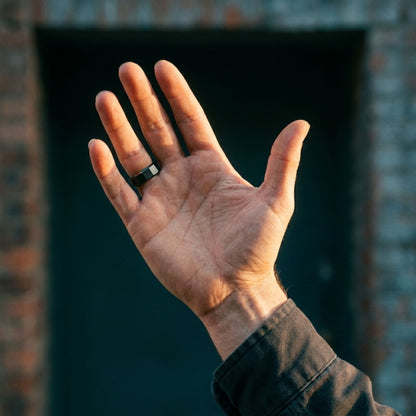 A close-up of a person's open hand wearing a simple black metal band ring, set against a blurred dark doorway and brick wall background.