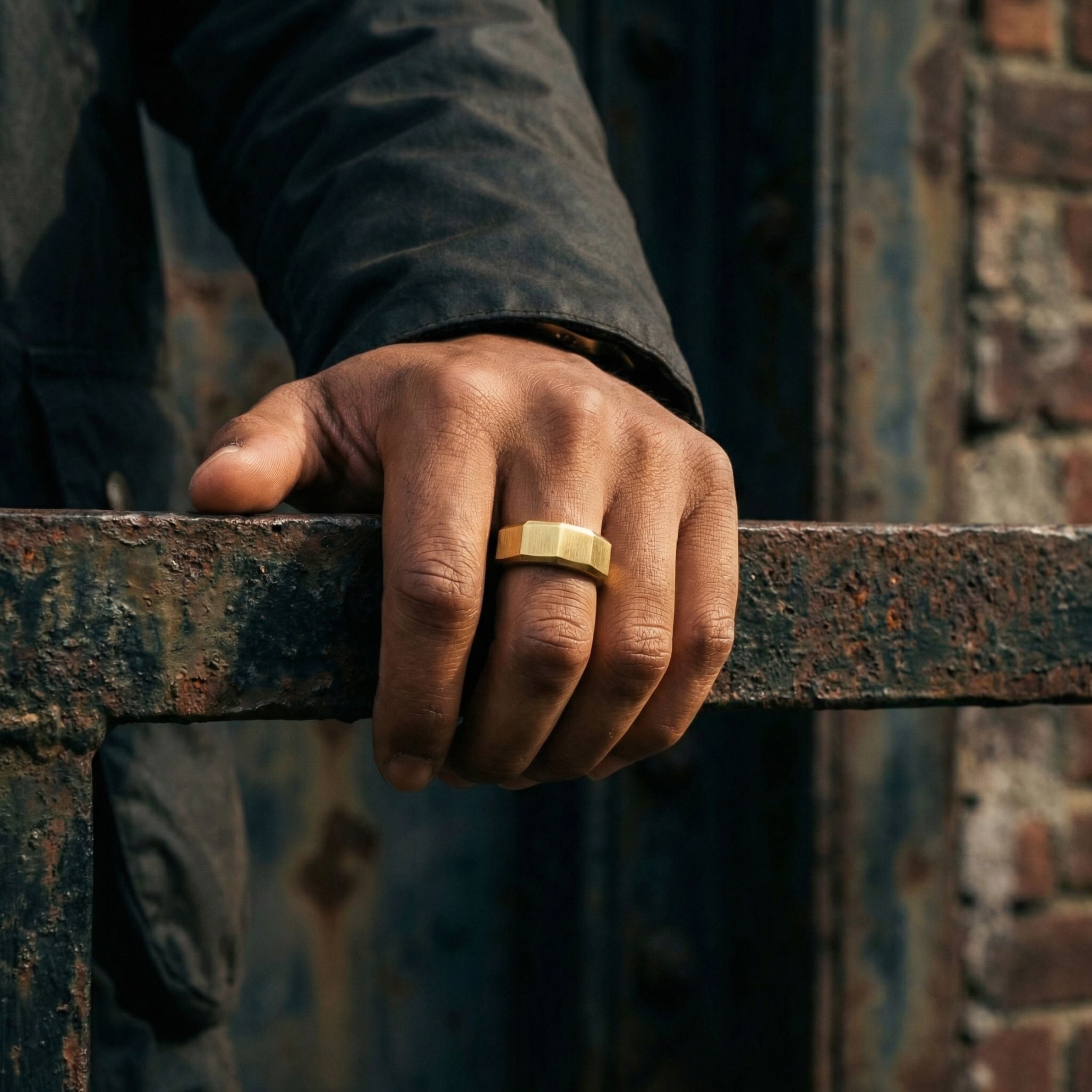 Gold stainless steel men's ring with a brushed geometric design worn on a hand resting on a weathered industrial steel railing