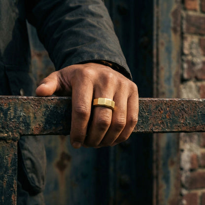 Gold stainless steel men's ring with a brushed geometric design worn on a hand resting on a weathered industrial steel railing