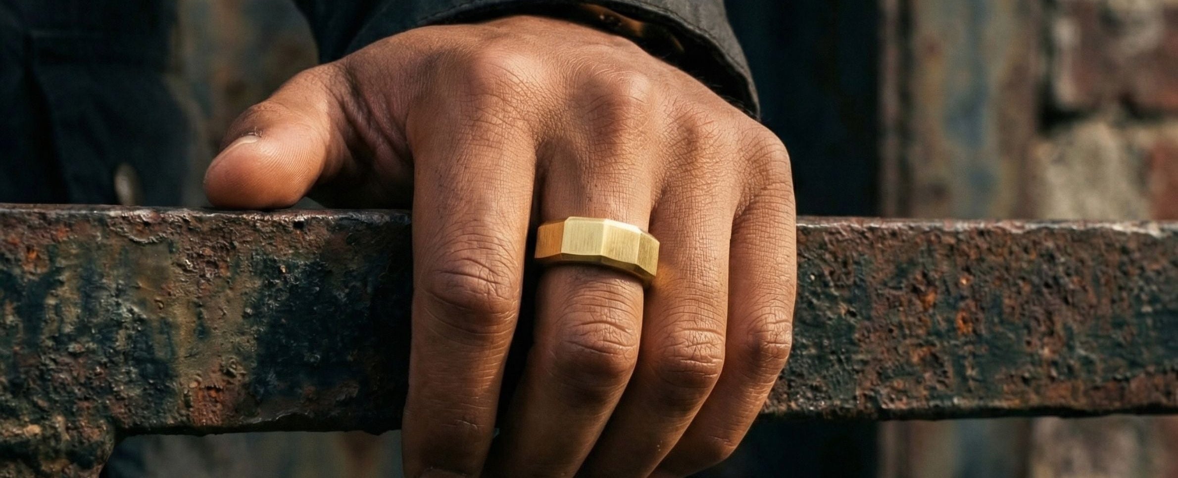 A close-up of a person's hand resting on a weathered metal railing, wearing a simple gold band ring. The background features a rustic, dark metal door and a brick wall.