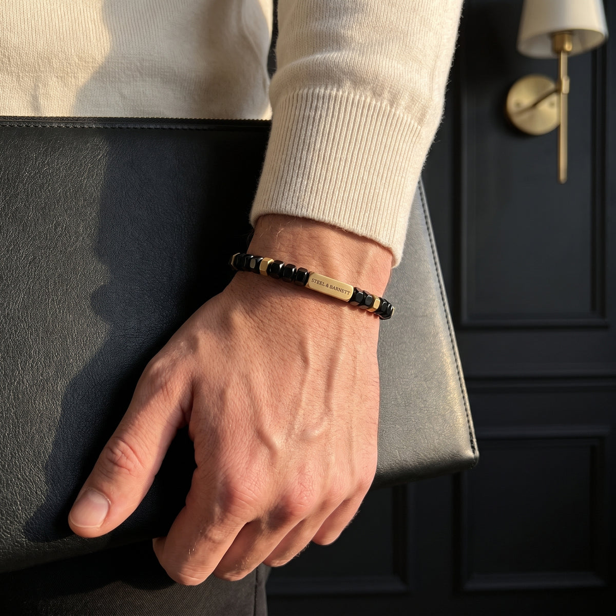 Black faceted bead men's bracelet with plated 18K gold clasp and accents worn on the wrist beside a black leather bag