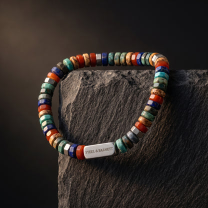 A beaded bracelet with a mix of colorful stone beads and a silver metal clasp engraved with 'STEEL & BARNETT', displayed on a dark textured rock against a dark background.