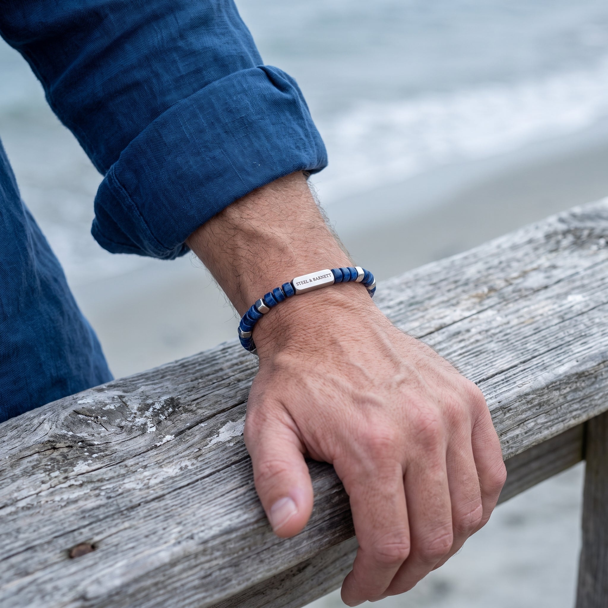 A close-up of a man's hand resting on a weathered wooden railing at the beach, wearing a beaded bracelet with blue and silver beads and a silver rectangular plate engraved.