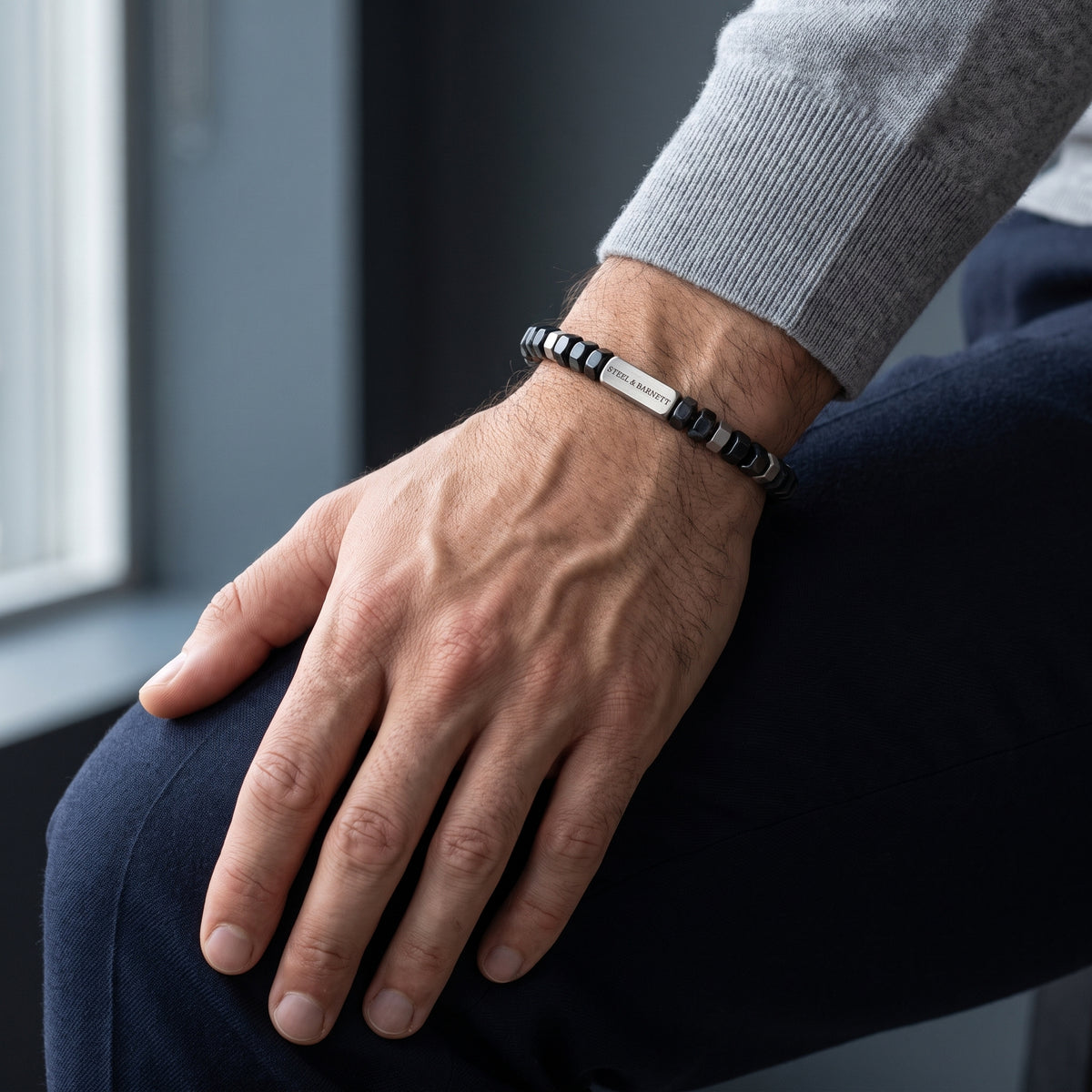 A close-up of a man's hand resting on his knee, wearing a beaded bracelet with a silver rectangular plate. The bracelet features alternating black and white beads, and the person is wearing a gray long-sleeve shirt and dark navy pants.
