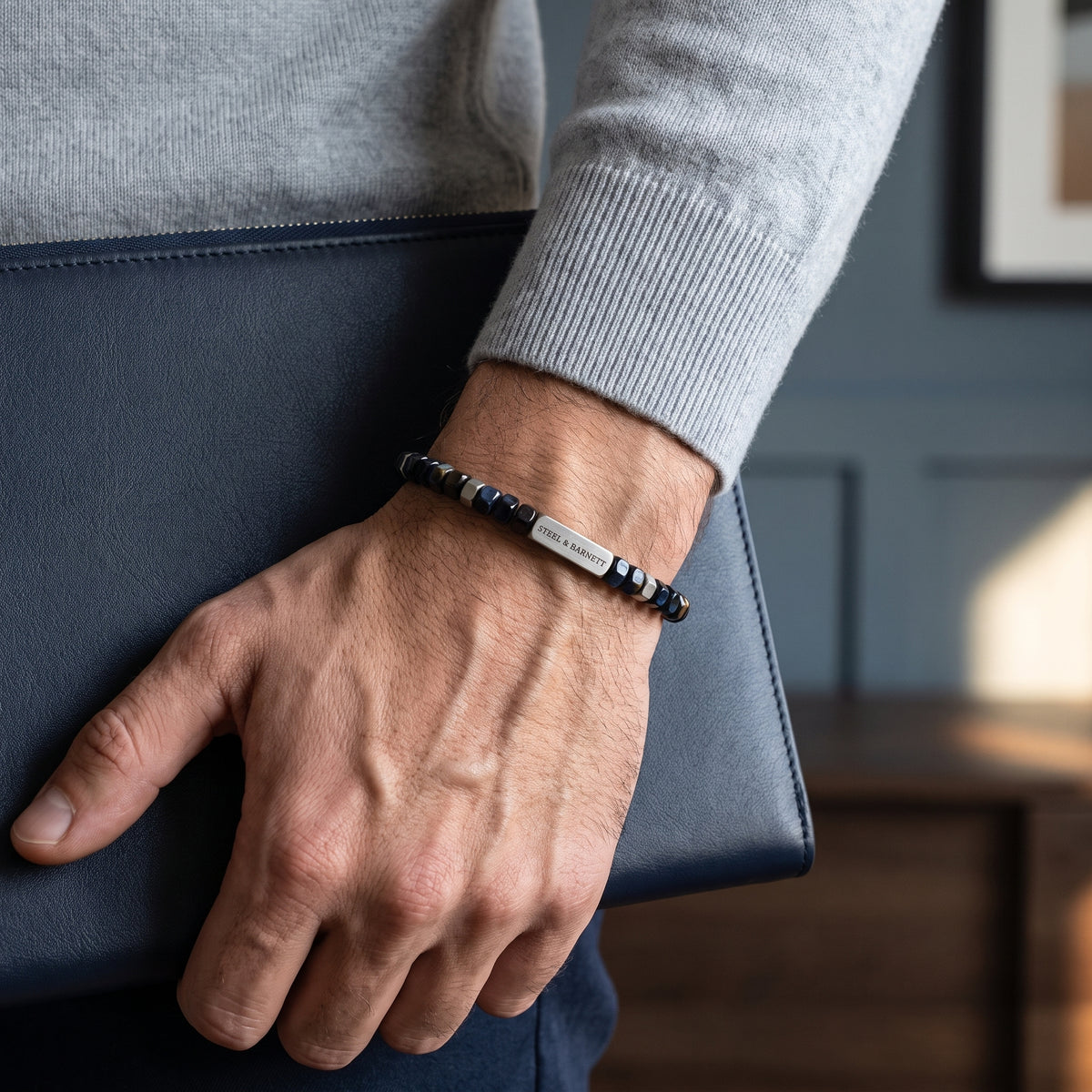 A close-up of a man's hand wearing a beaded bracelet with a silver-toned rectangular plate, holding a dark blue leather folder. He is wearing a light gray knit sweater, and the background shows a softly blurred blue-gray wall and wooden furniture.