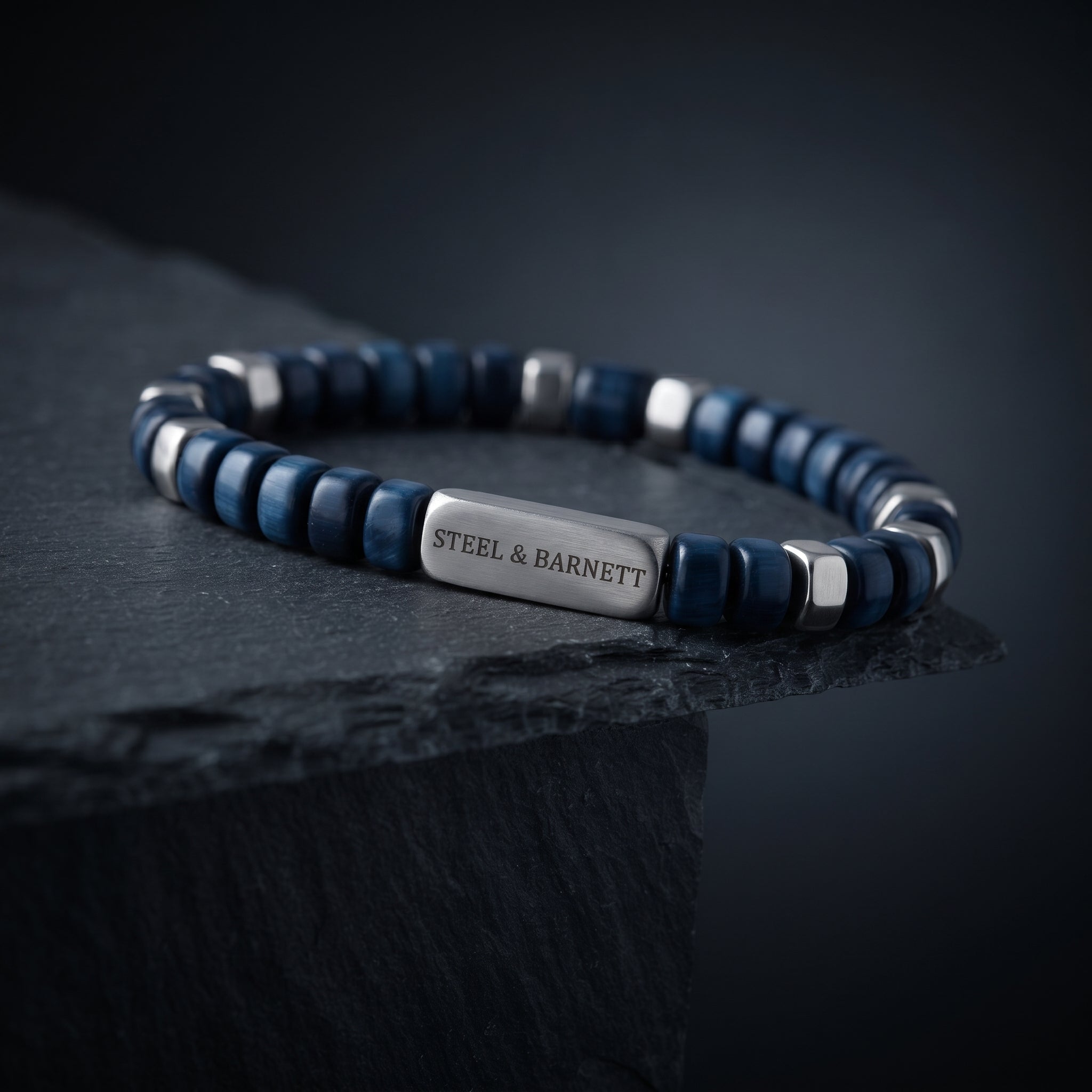 A beaded stretch bracelet with dark blue and silver beads, featuring a central silver plate engraved with 'STEEL & BARNETT', displayed on a dark slate surface.