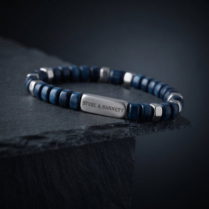 A beaded stretch bracelet with dark blue and silver beads, featuring a central silver plate engraved with 'STEEL & BARNETT', displayed on a dark slate surface.
