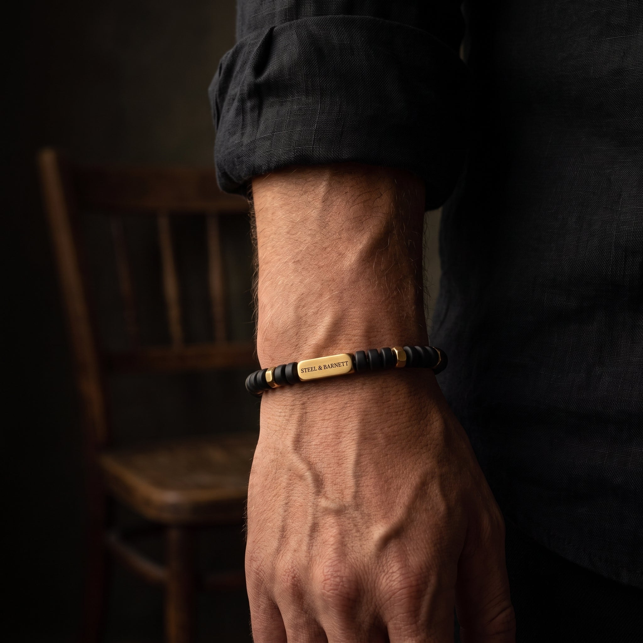 A close-up of a man's hand and wrist wearing a beaded bracelet with a central gold-toned metal plate engraved with 'STEEL & BARNETT'. The background is dark and moody with a wooden chair visible.