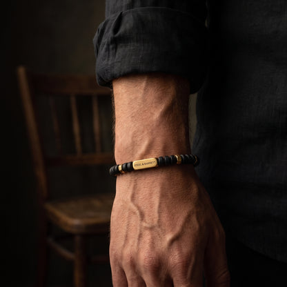 A close-up of a man's hand and wrist wearing a beaded bracelet with a central gold-toned metal plate engraved with 'STEEL & BARNETT'. The background is dark and moody with a wooden chair visible.