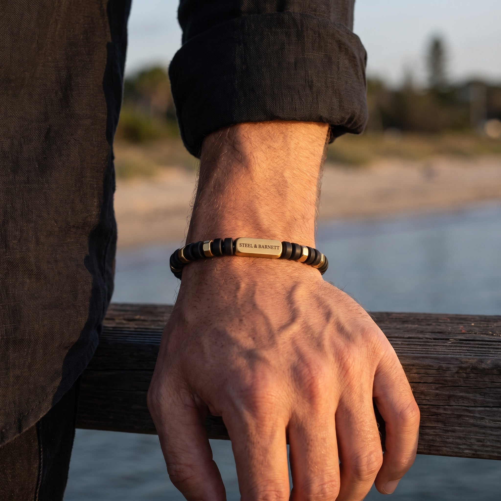 A close-up of a man's hand resting on a wooden railing by the water, wearing a personalized beaded bracelet with a gold-toned central plate engraved with 'STEEL & BARNETT'.