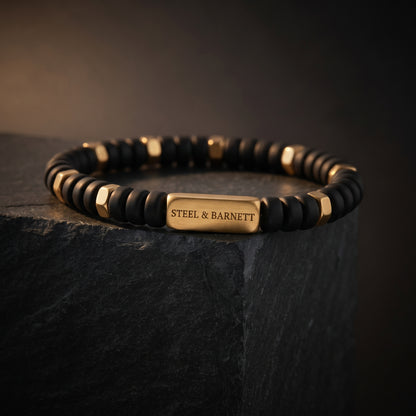 A personalized beaded bracelet with matte black round beads, faceted gold spacer beads, and a central gold rectangular plate engraved with 'STEEL & BARNETT', displayed on a dark slate pedestal.