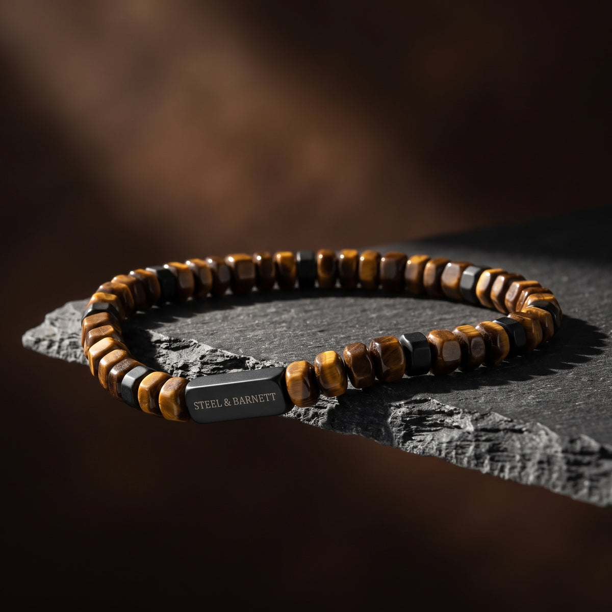 A beaded bracelet with alternating brown and black tiger's eye beads and a rectangular steel clasp engraved with 'STEEL & BARNETT', resting on a dark textured stone surface against a dark brown background.