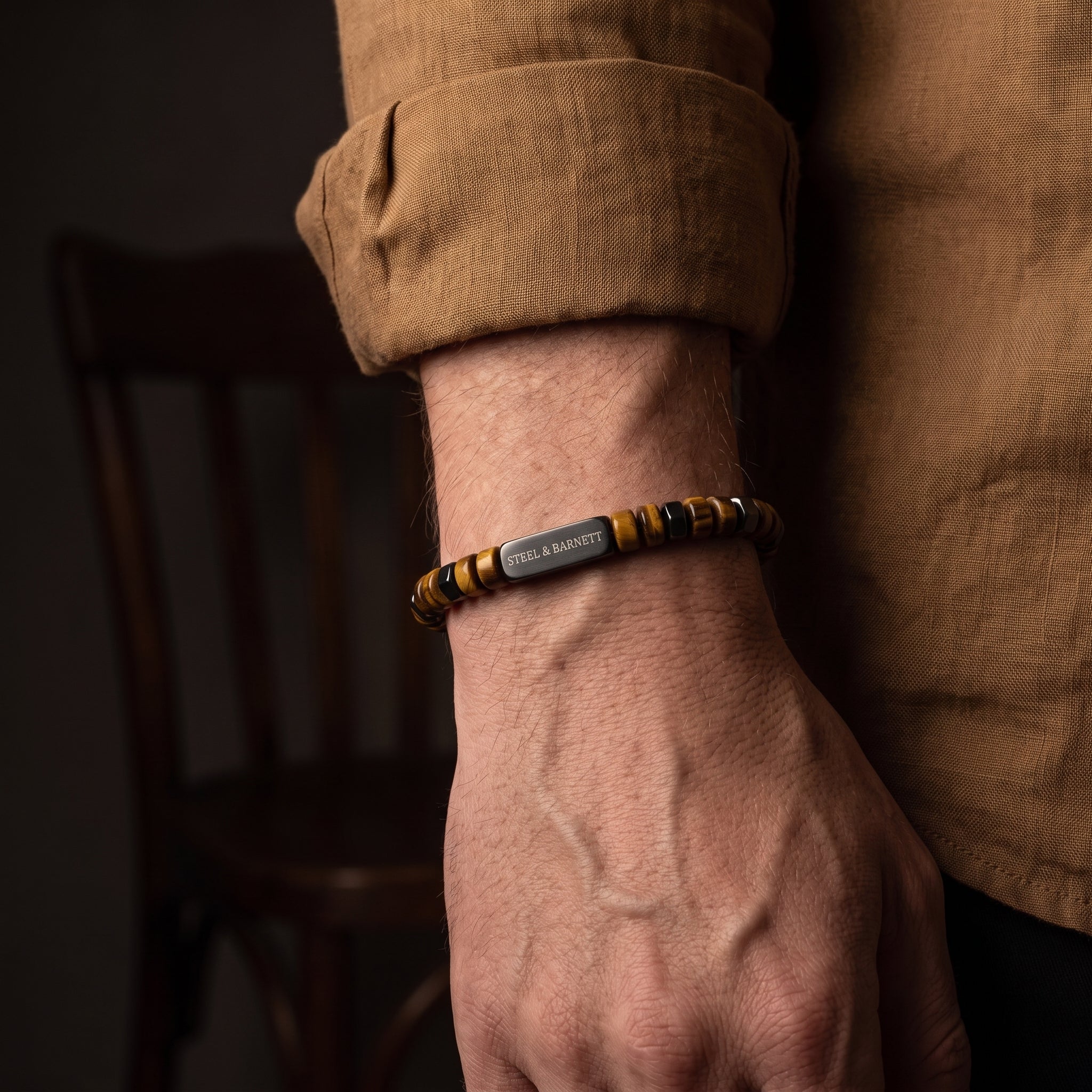 A close-up of a man's wrist wearing a beaded bracelet with a central silver-toned metal plate engraved with 'STEEL & BARNETT'. The bracelet features alternating dark brown and yellow-gold beads, paired with black and gold spacer beads.