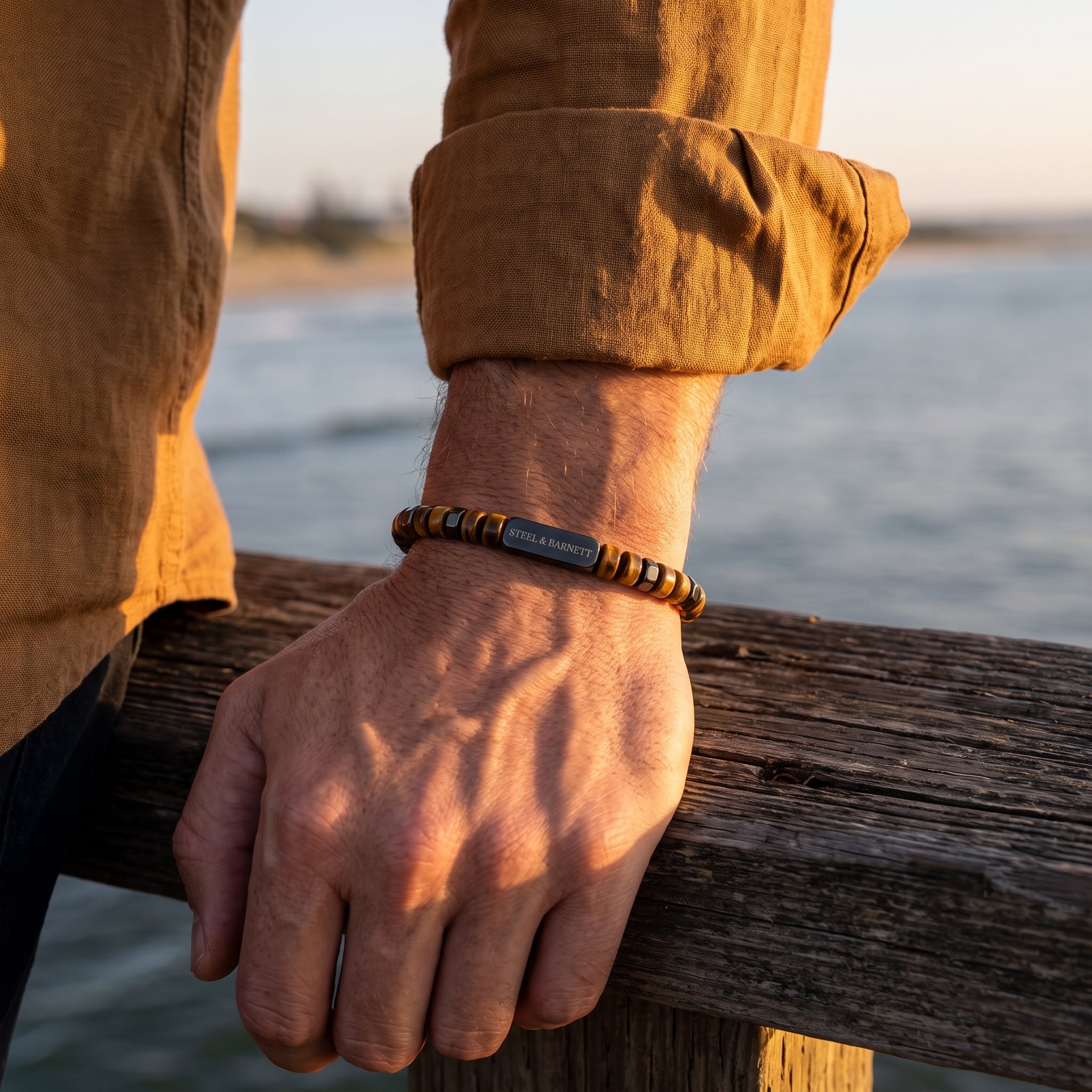 A close-up of a man's hand resting on a wooden railing outdoors near the water, wearing a beaded bracelet with a central gray metal tag engraved with 'STEEL & BARNETT'.
