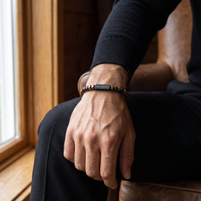 A close-up of a man's hand resting on his knee, wearing a beaded bracelet with a central black rectangular plate engraved with the brand name 'STEEL & BARNETT'.