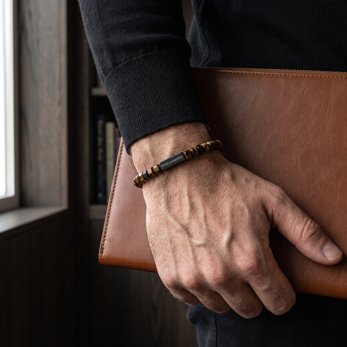 Brown and black faceted bead men's bracelet with a black branded bar worn on the wrist beside a brown leather portfolio