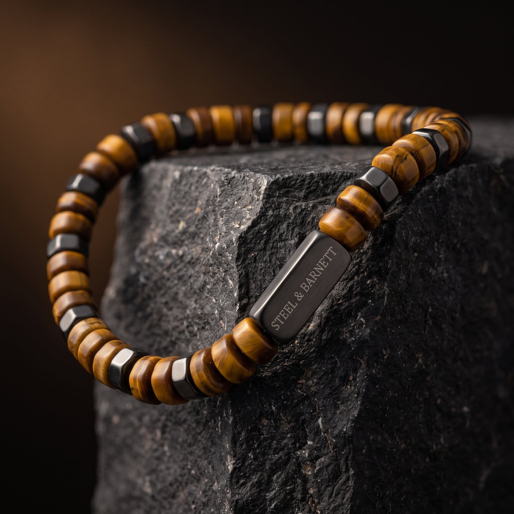 A beaded bracelet with alternating brown tiger's eye and black ceramic beads, featuring a polished black metal clasp engraved with 'STEEL & BARNETT'. The bracelet is displayed on a dark textured rock against a dark, warm-toned background.