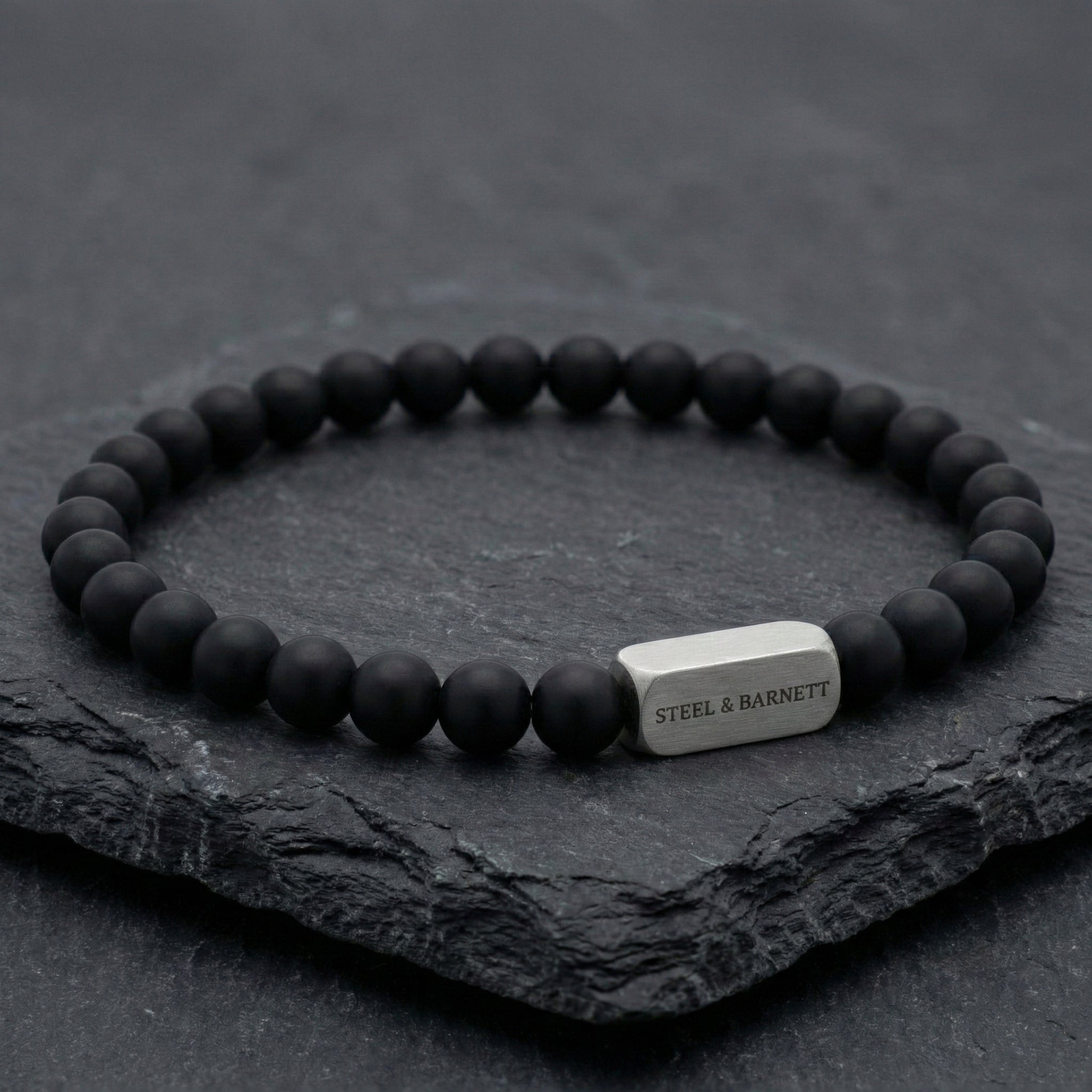black beaded men’s bracelet with brushed stainless steel bar detail on dark stone surface