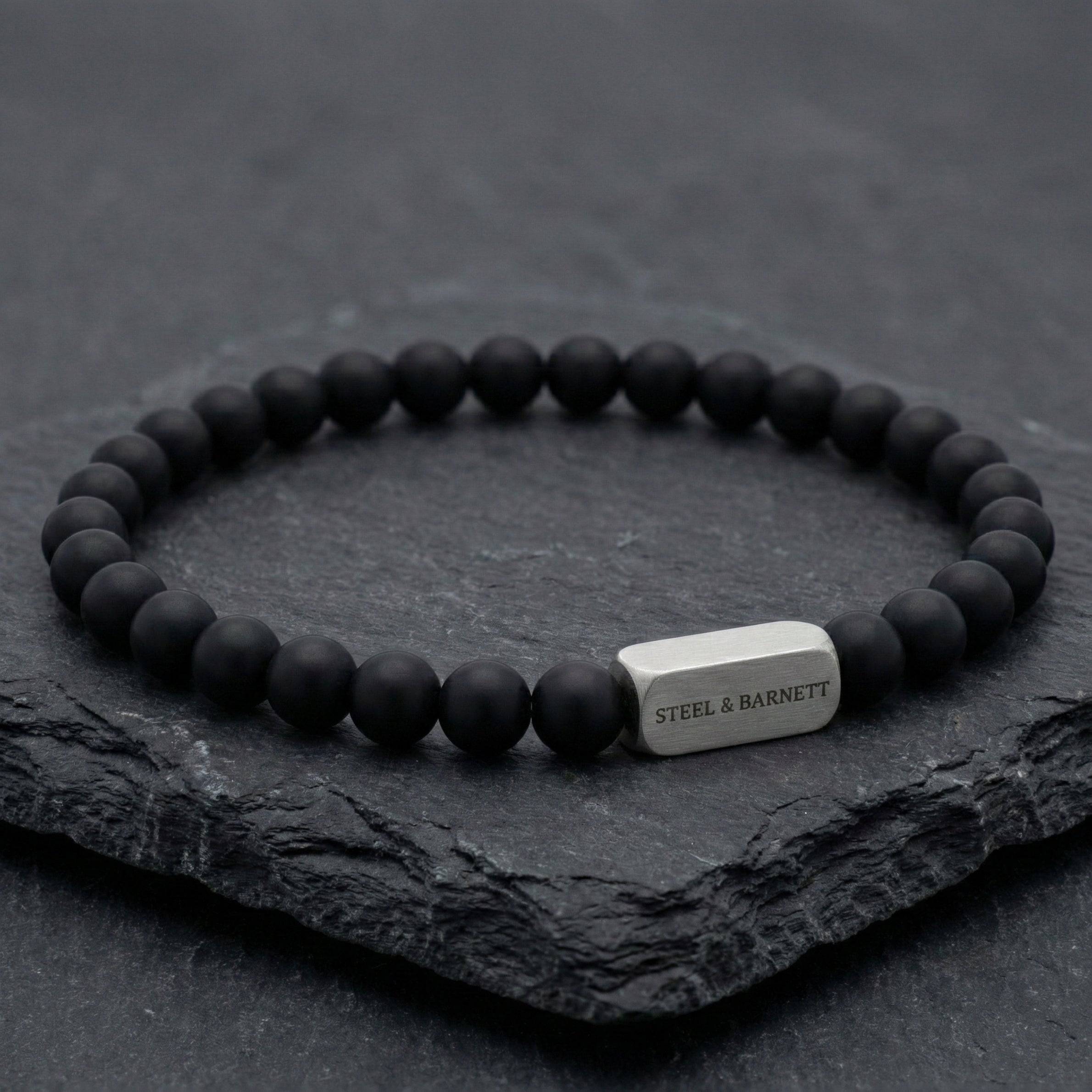 black beaded men’s bracelet with brushed stainless steel bar detail on dark stone surface