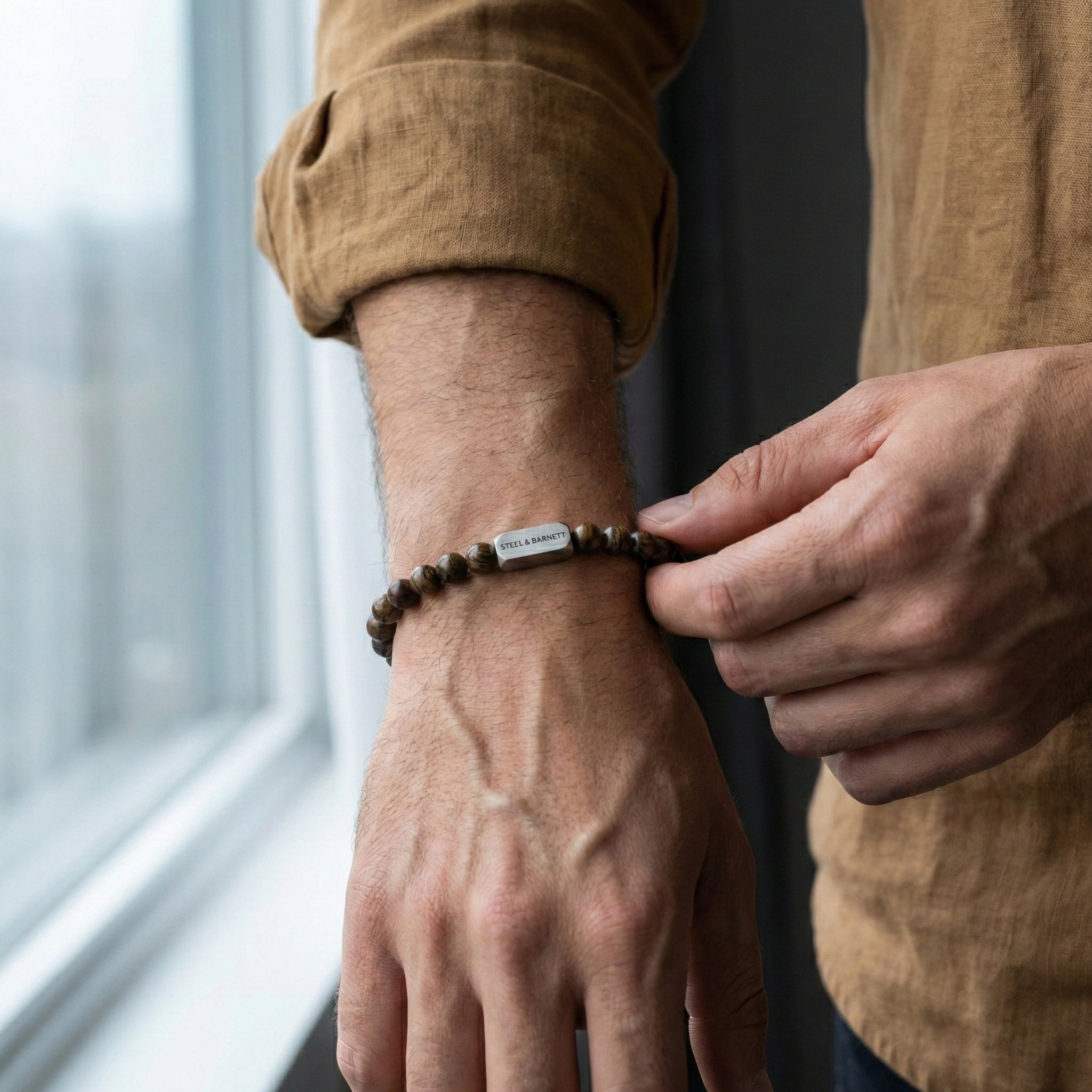 Bronzite beaded men’s bracelet with brown stone beads and brushed stainless steel bar worn on wrist by window