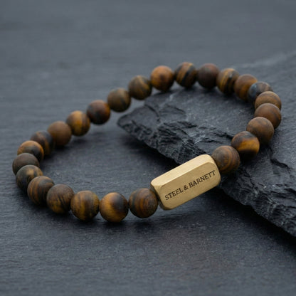 Tiger eye men’s bracelet with round natural stone beads and plated 18K gold clasp on dark stone surface