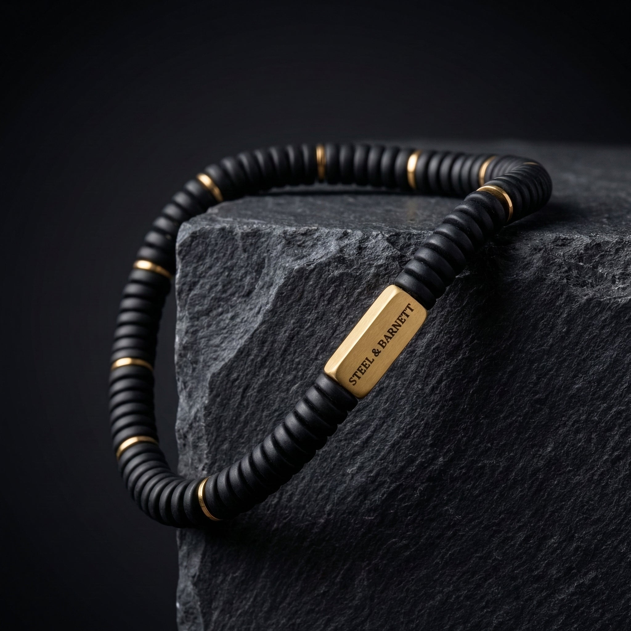 Black beaded men's bracelet with matte disc beads and rectangular plated 18K gold accent plate displayed on a dark stone block close up detail