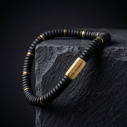 Black beaded men's bracelet with matte disc beads and rectangular plated 18K gold accent plate displayed on a dark stone block close up detail