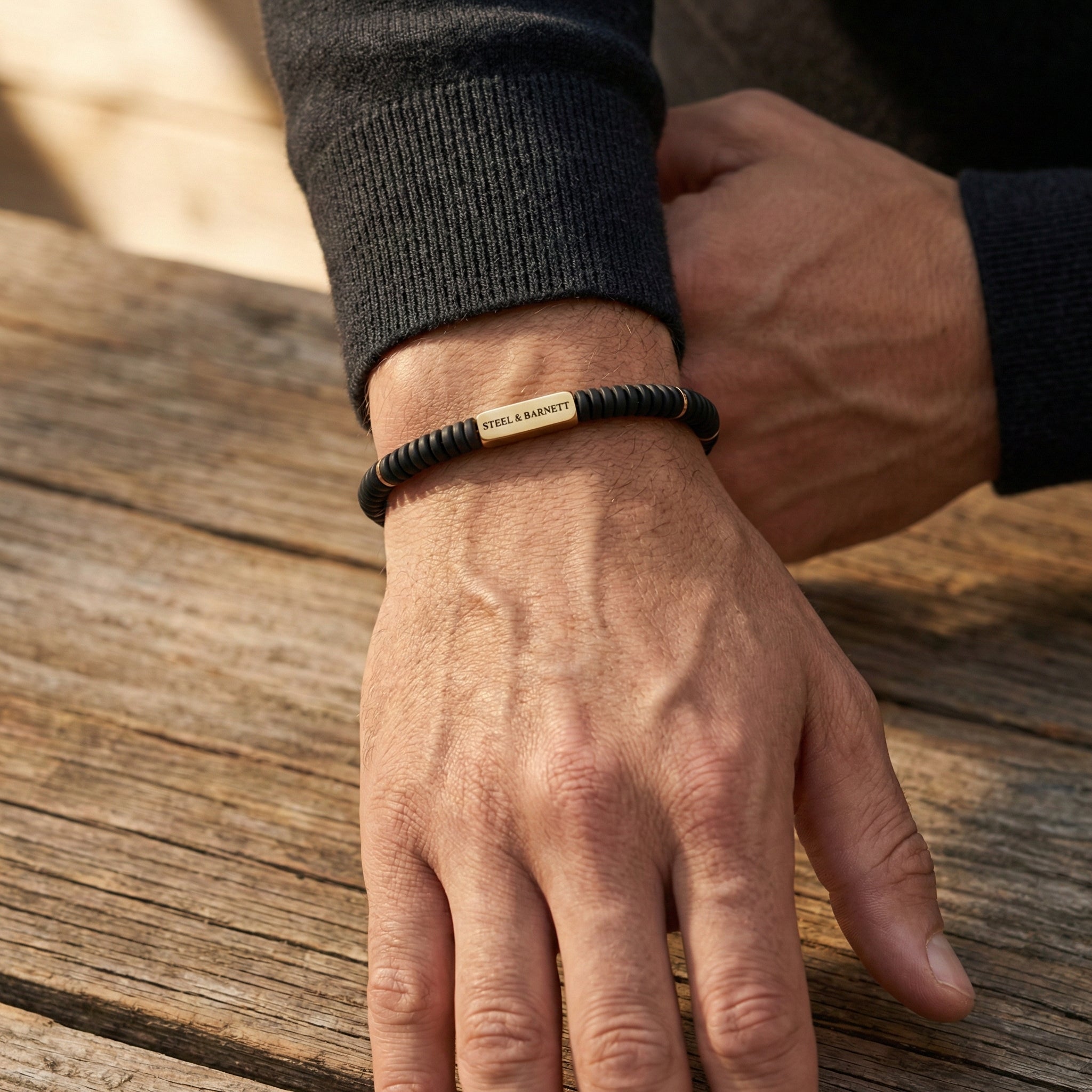 Black beaded men's bracelet with matte disc beads and rectangular plated 18K gold accent plate worn on wrist above a wooden surface