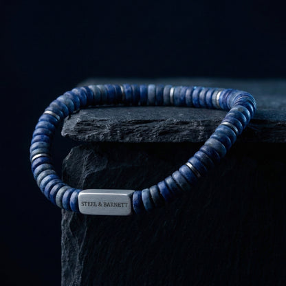 Blue natural stone men's beaded bracelet with brushed stainless steel accent plate displayed on a dark stone block close up detail