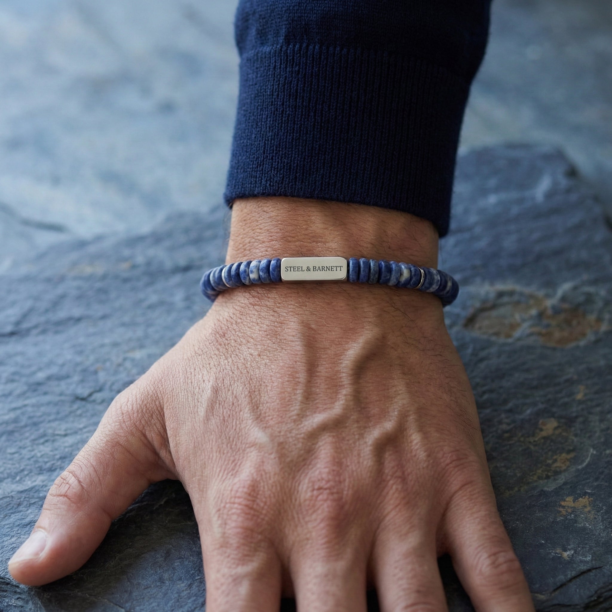 Blue natural stone men's beaded bracelet with brushed stainless steel accent plate worn on wrist on a stone surface