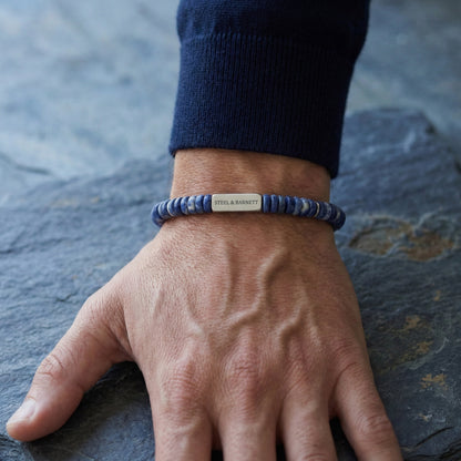 Blue natural stone men's beaded bracelet with brushed stainless steel accent plate worn on wrist on a stone surface