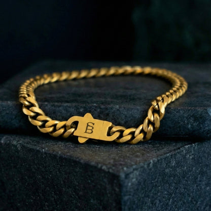 Gold toned stainless steel men’s bracelet with Cuban chain links and plated 18K gold clasp on dark stone surface close up