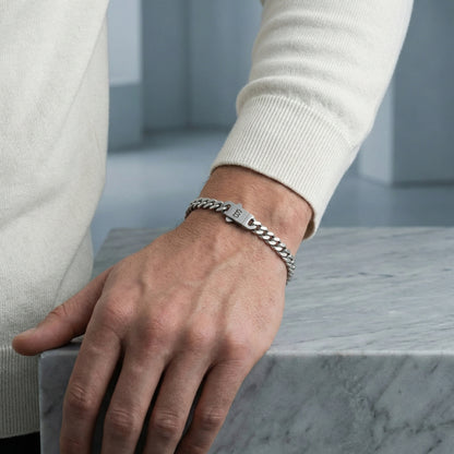 Silver toned stainless steel men’s bracelet with Cuban chain links and engraved clasp shown on wrist resting on marble surface