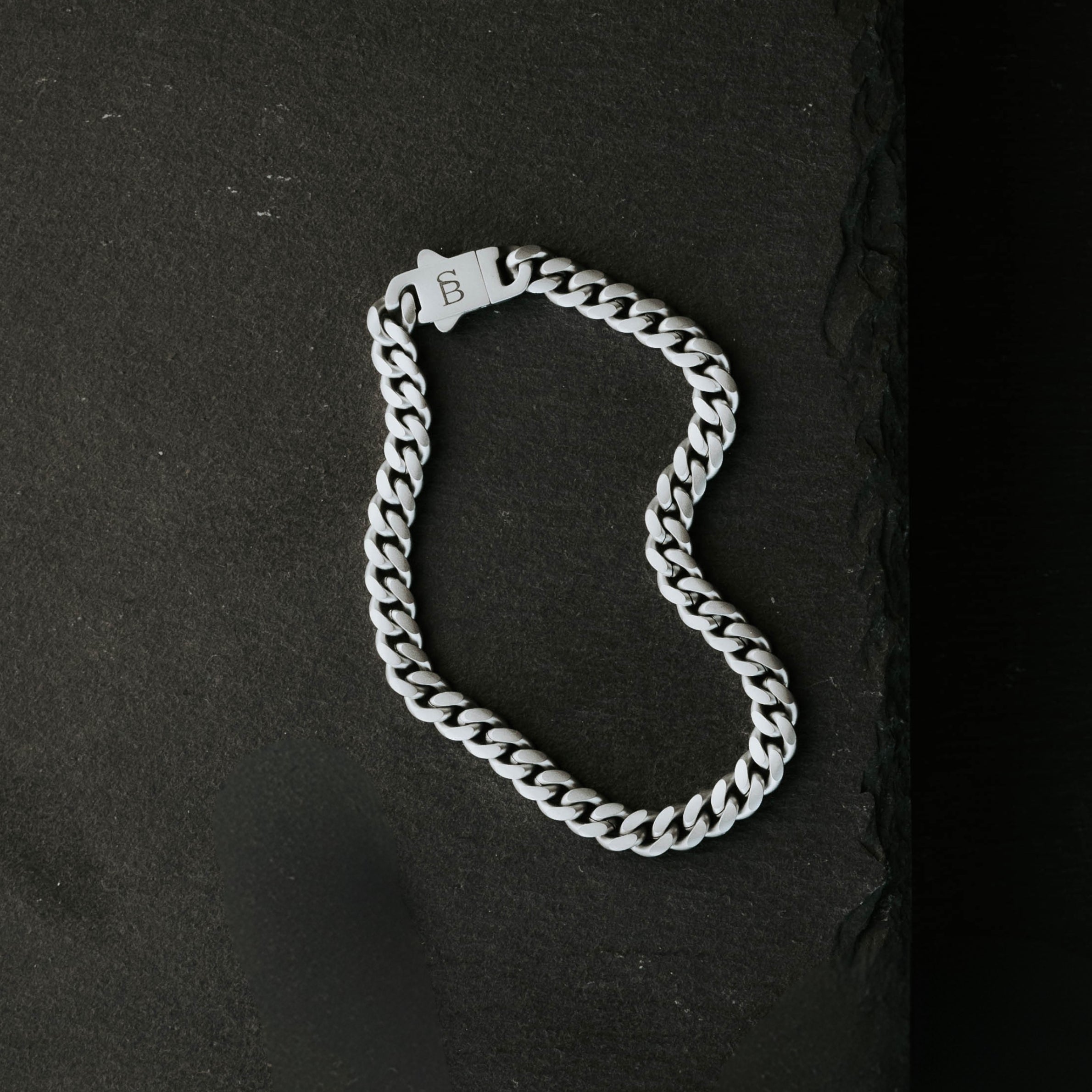 Silver toned stainless steel men’s bracelet with Cuban chain links and engraved clasp on dark stone surface