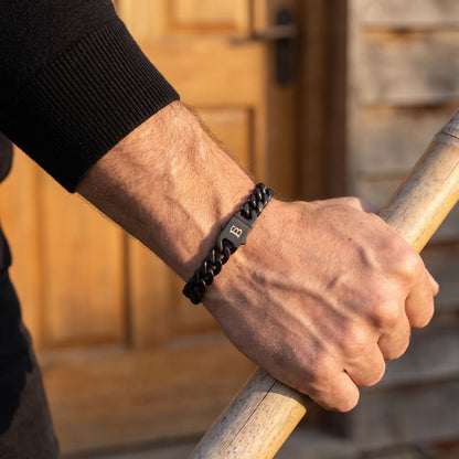 Black stainless steel men's bracelet with a curb chain design and engraved clasp worn on a wrist while holding a wooden handle