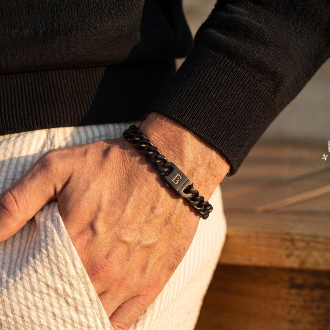 Black stainless steel men's bracelet with a curb chain design and engraved clasp worn on a wrist in an outdoor setting