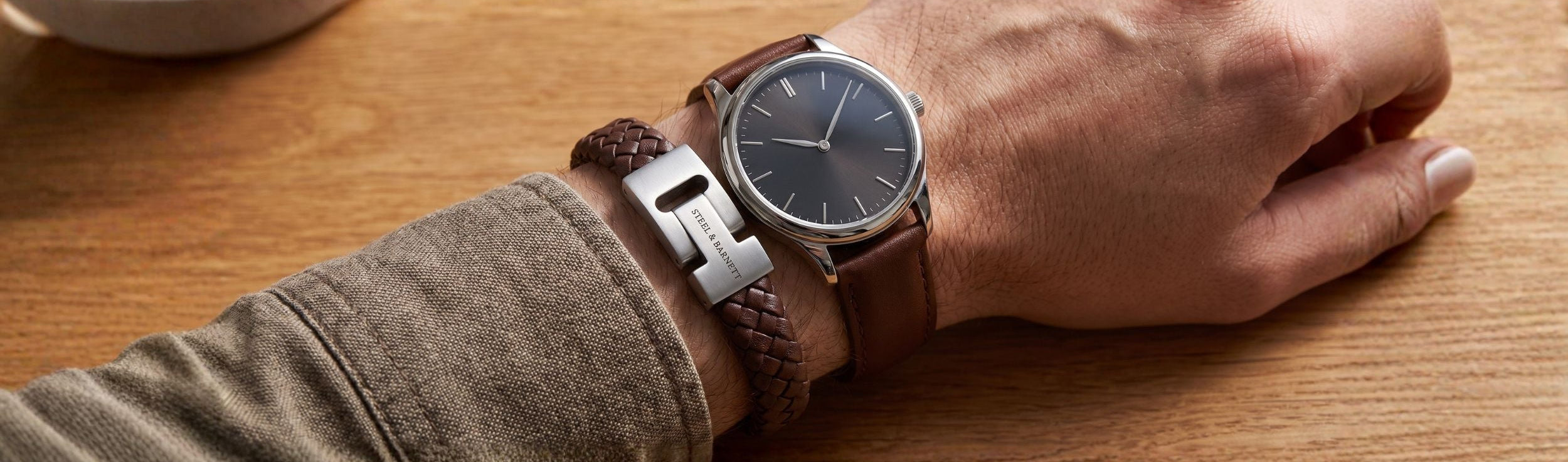 A close-up of a person's wrist wearing a round analog watch with a dark dial, brown leather strap, and silver-toned buckle, resting on a wooden table.