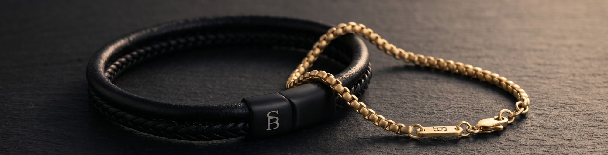 A black leather bracelet with a gold chain link section and a black magnetic clasp featuring a white monogram 'B', displayed on a dark slate surface.