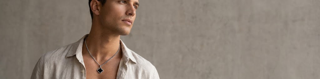 A man wearing a silver-toned necklace with a black clover-shaped pendant, styled with an open-collar light beige shirt against a textured gray concrete wall.