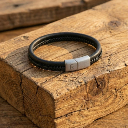 Black cowhide leather bracelet with braided band and brushed stainless steel magnetic clasp resting on wooden surface