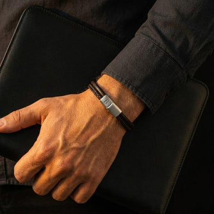 Brown leather men's bracelet with braided detail and stainless steel magnetic clasp worn on wrist holding black leather notebook