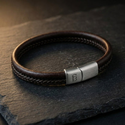 Brown leather men's bracelet with braided center strand and brushed stainless steel magnetic clasp shown in close up on dark stone surface
